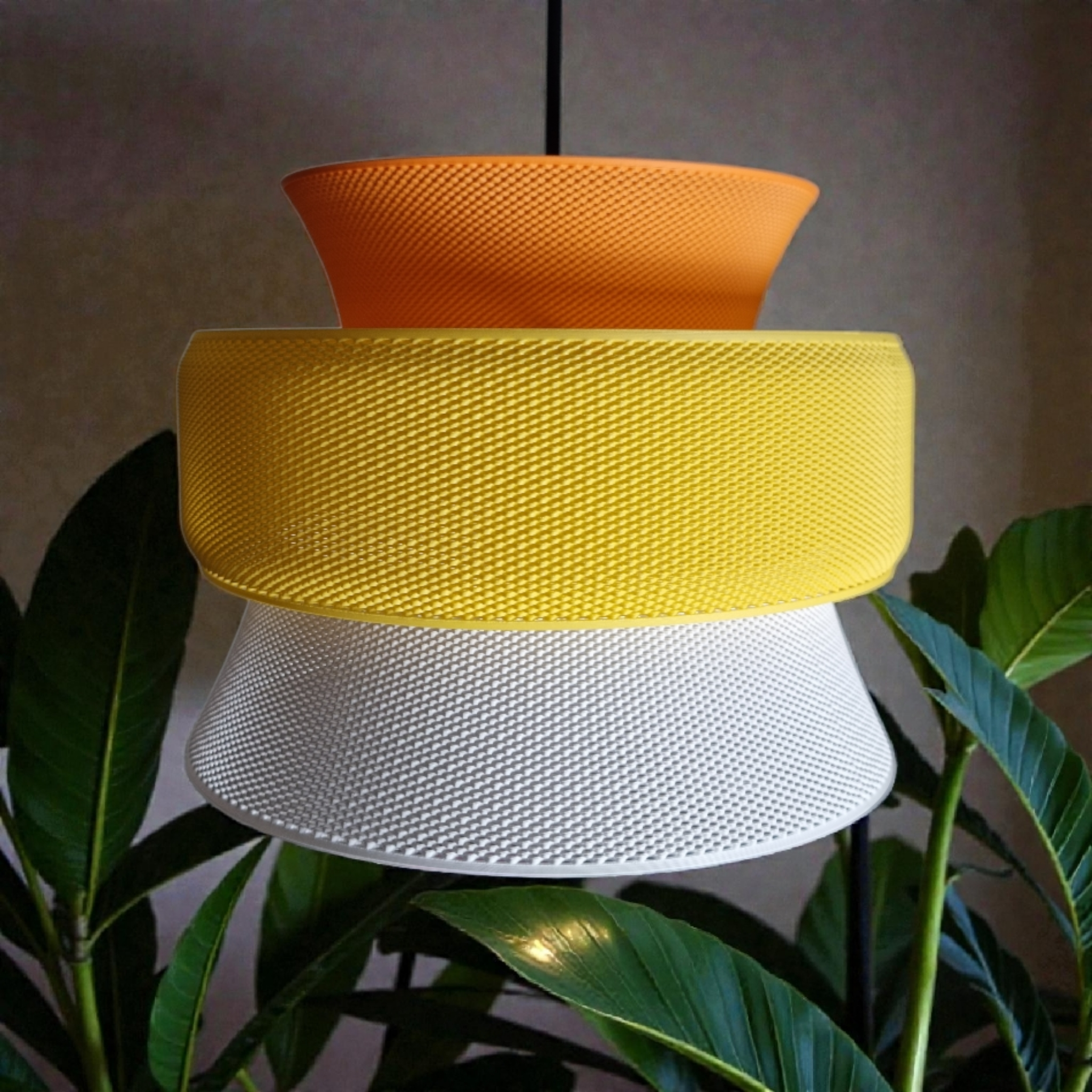 A modern pendant light features three stacked, textured segments in vibrant orange, yellow, and white, set against a backdrop of lush green plants. The geometric design enhances light diffusion, creating an eye-catching focal point.