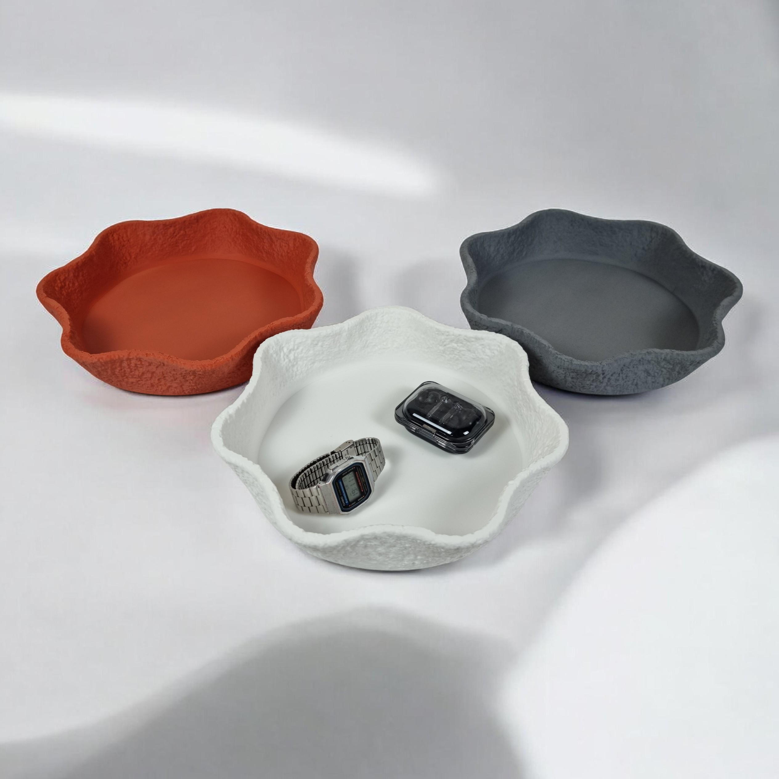 Three decorative bowls in varying colors—rusty red, white, and dark gray—are arranged on a light backdrop. Each bowl features an irregular, textured edge and holds a watch and a bracelet, showcasing their stylish functionality.