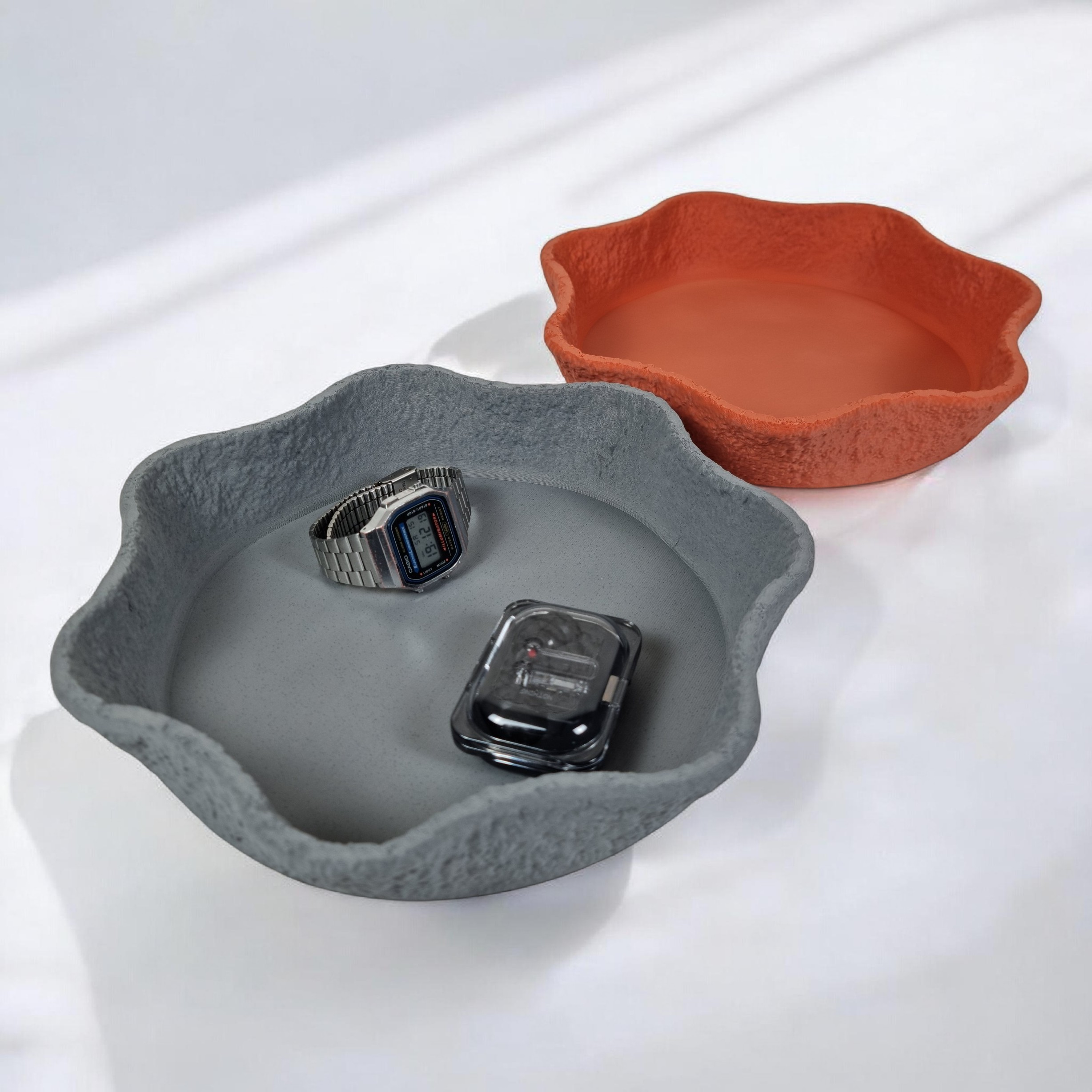 Two decorative bowls are displayed side by side on a bright surface. The left bowl is a textured gray with irregular edges, while the right bowl is a smooth orange, both holding small items like a watch and a gadget.