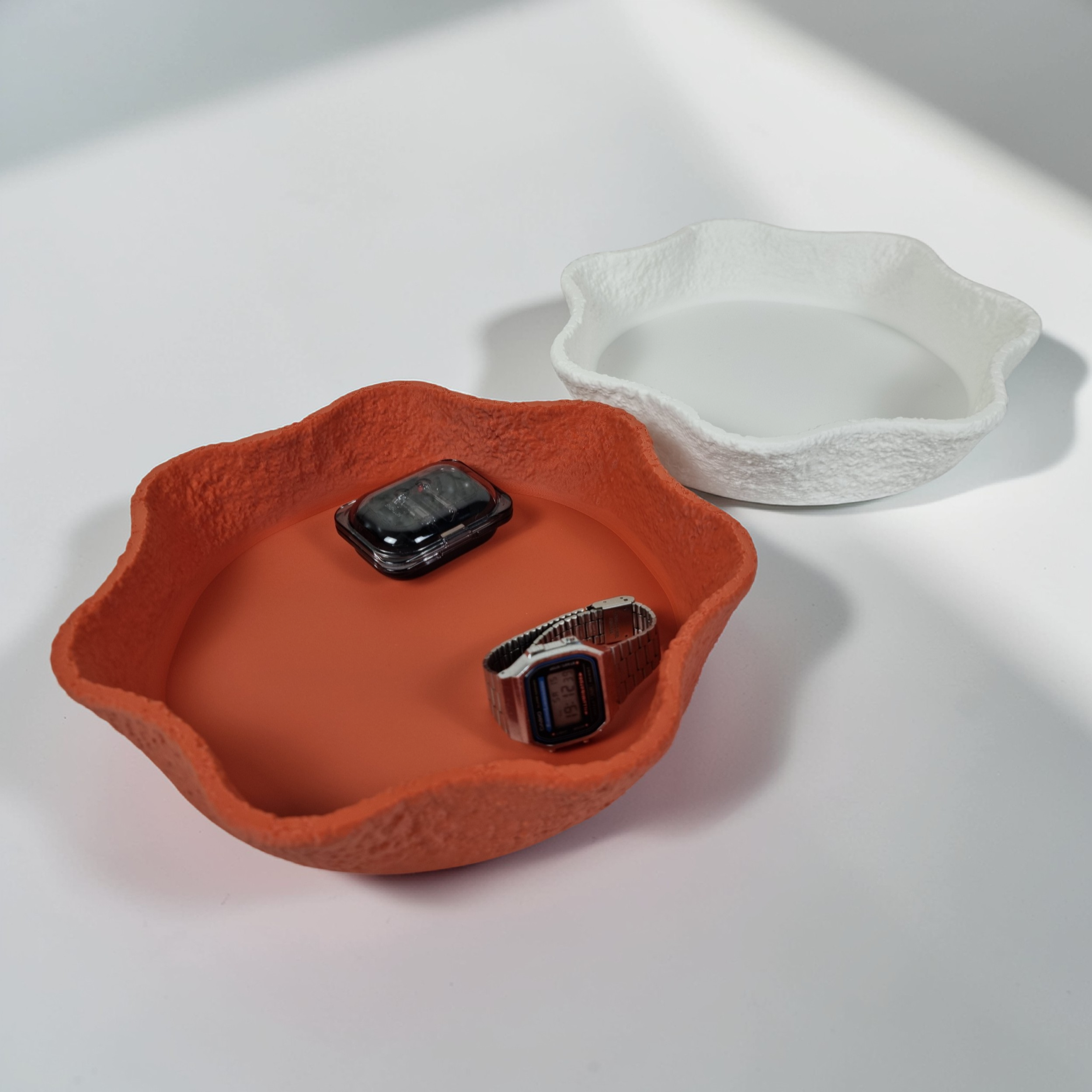 Two decorative bowls are shown side by side on a light surface. The bowl on the left is textured and orange with irregular edges, while the bowl on the right is smooth and white, both containing small items like watches.