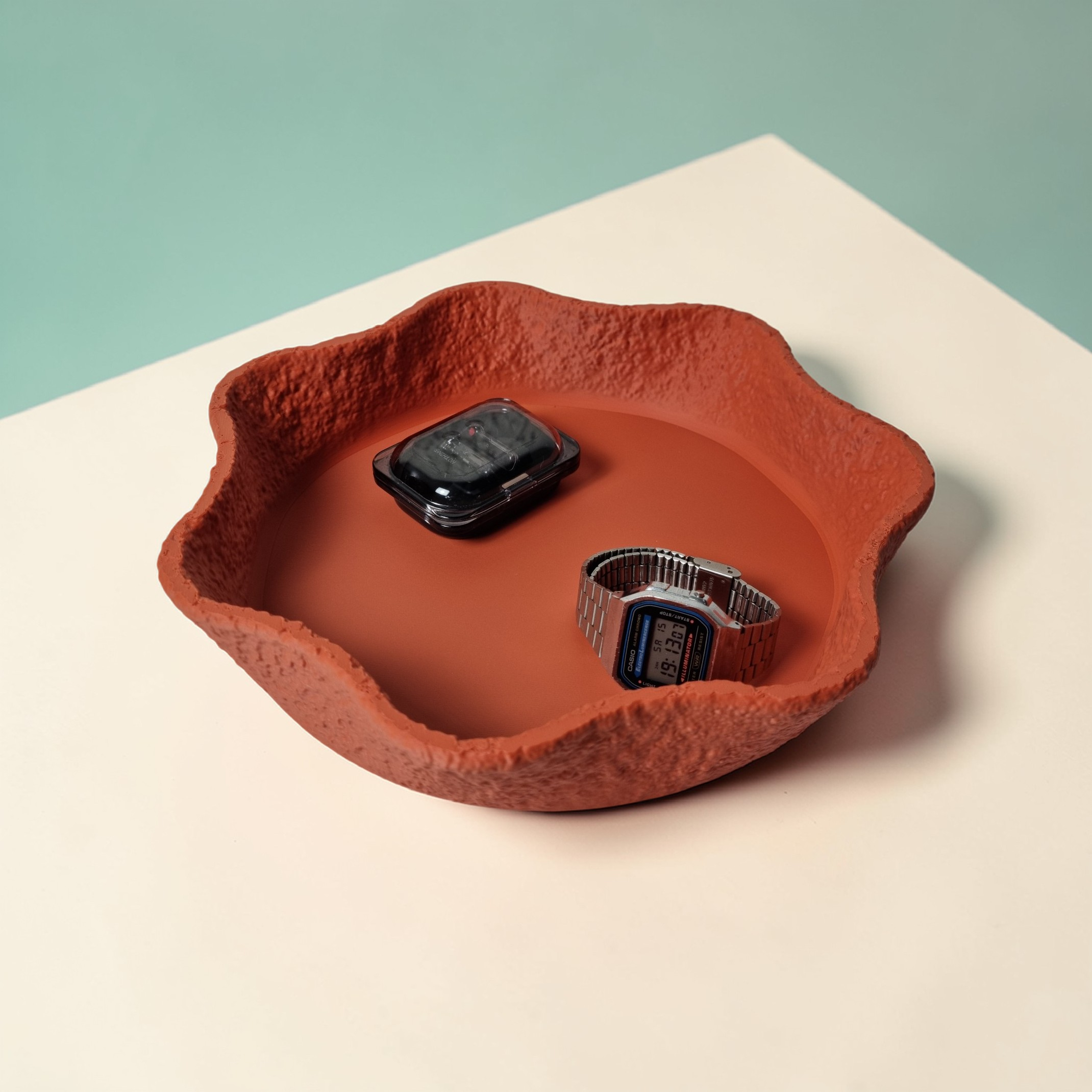 A textured, organic-shaped clay dish in a warm terracotta color sits on a light beige surface. Inside, it holds two watches—one digital and one analog—against a smooth, contrasting base, showcasing a blend of rustic and modern design.