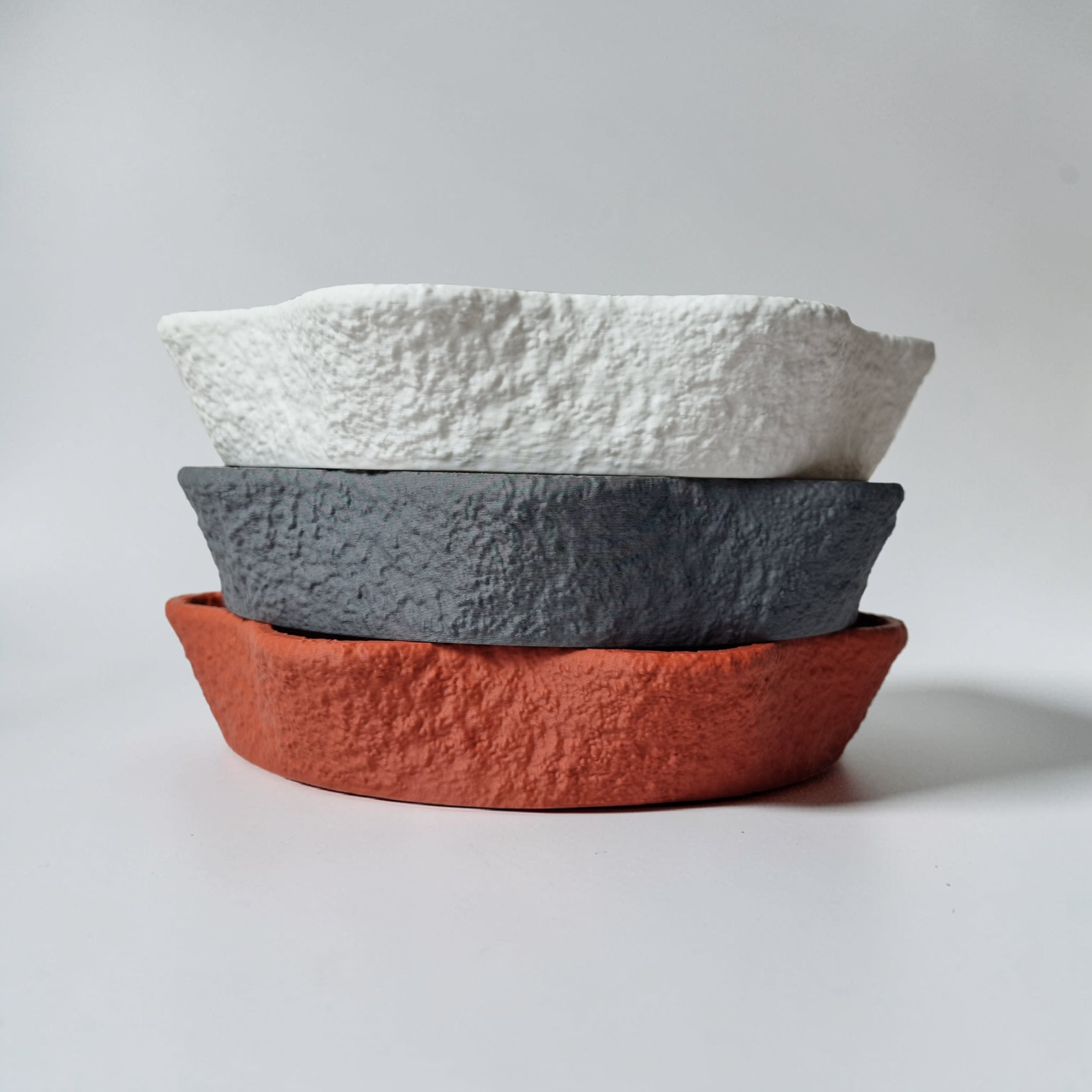 Three decorative bowls are stacked on top of each other, showcasing a textured, organic design. The top bowl is white, the middle one is gray, and the bottom one is a warm terracotta color, all set against a plain white background.