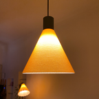 A modern pendant lamp with a conical shape features a textured orange shade that emits a warm glow, illuminating the surrounding space. In the background, a similar lamp with a white shade adds to the cozy ambiance.