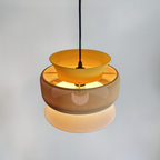 SAN pendant lamp switched on, casting warm light through its three layered rings in textured bioplastic — mustard yellow, sand and white