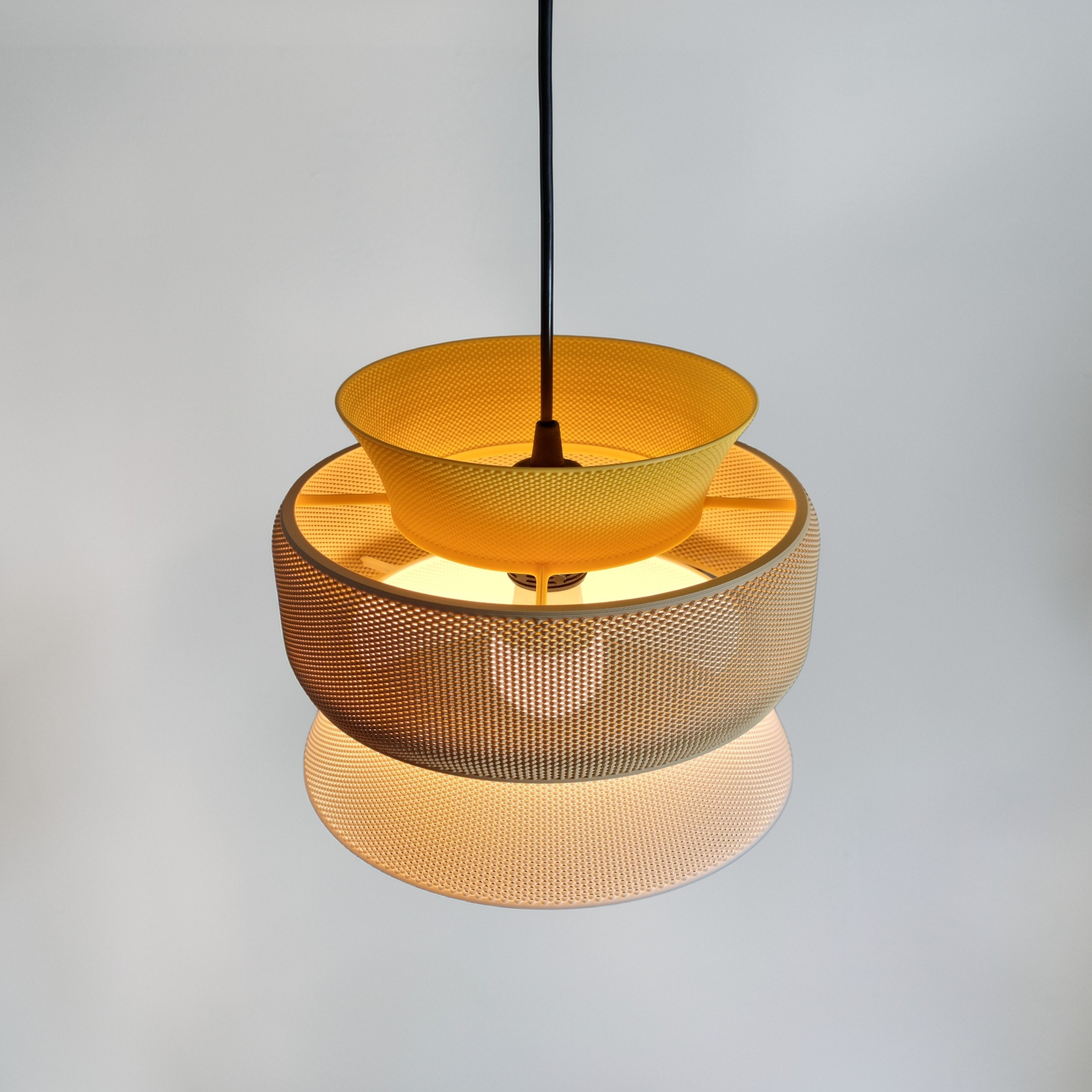 SAN pendant lamp switched on, casting warm light through its three layered rings in textured bioplastic — mustard yellow, sand and white