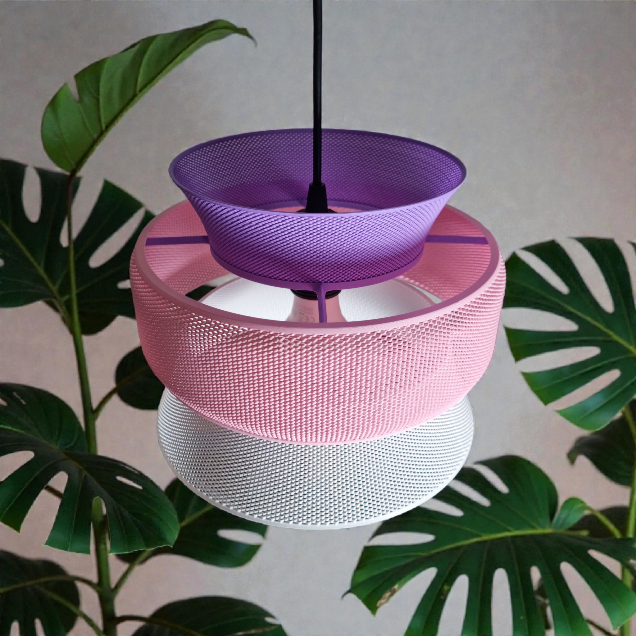 A colorful pendant lamp featuring three stacked mesh segments in purple, pink, and white, suspended from a black cord. The lamp is set against a backdrop of large green monstera leaves, creating a vibrant, modern aesthetic.