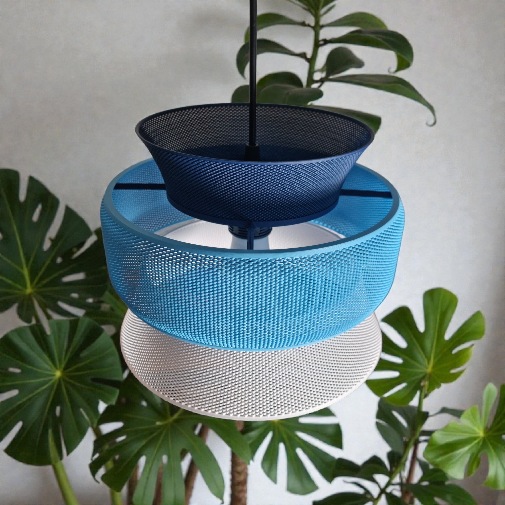 A modern pendant light featuring three distinct mesh segments in blue, navy, and white, suspended from a black cord. The light is set against a backdrop of lush green plants, creating a stylish and contemporary atmosphere.