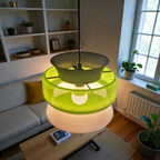 A modern pendant lamp features a layered design with three distinct segments in green, white, and a warm glow, suspended from a black cord. The lamp illuminates a cozy living space with plants and a light-colored sofa in the background.