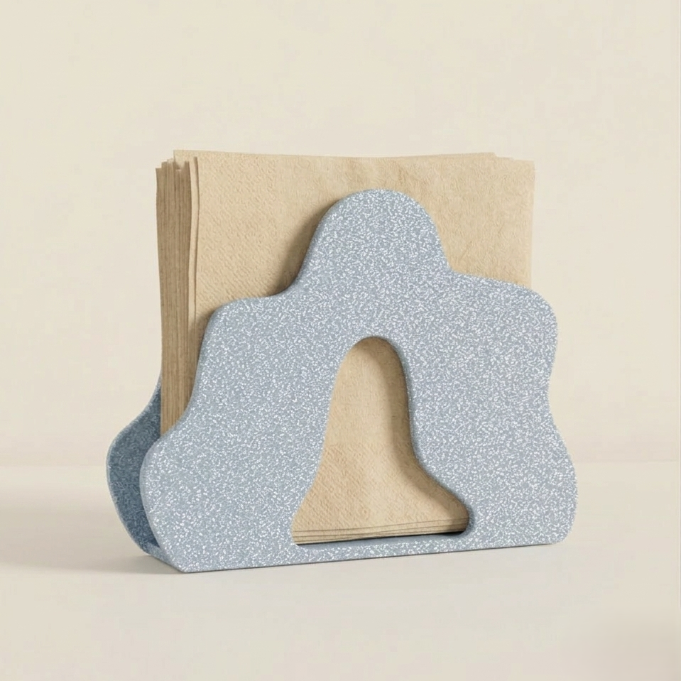 A minimalist napkin holder in a soft blue color with a textured finish, featuring an organic, rounded shape. It holds neatly stacked beige napkins, adding a contemporary touch to any dining or kitchen space.