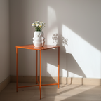 A white sculptural vase resembling a small ghost sits atop a vibrant orange side table. The vase is filled with a bouquet of daisies, casting soft shadows on the light-colored wall as sunlight streams.