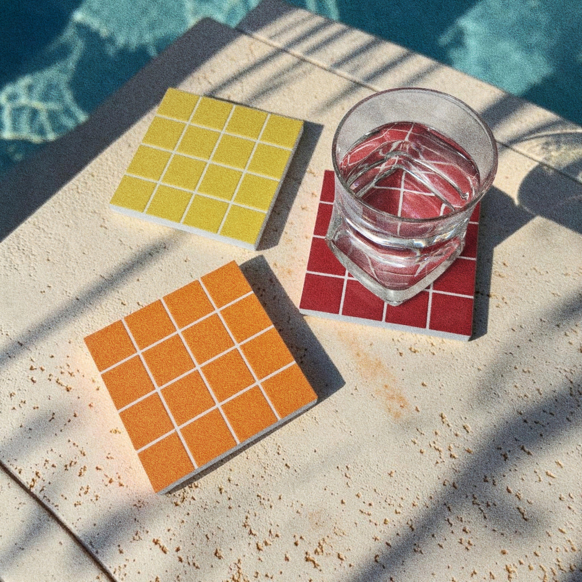 Three TILES coasters by a swimming pool, a glass of water resting on the red one in sunlight
