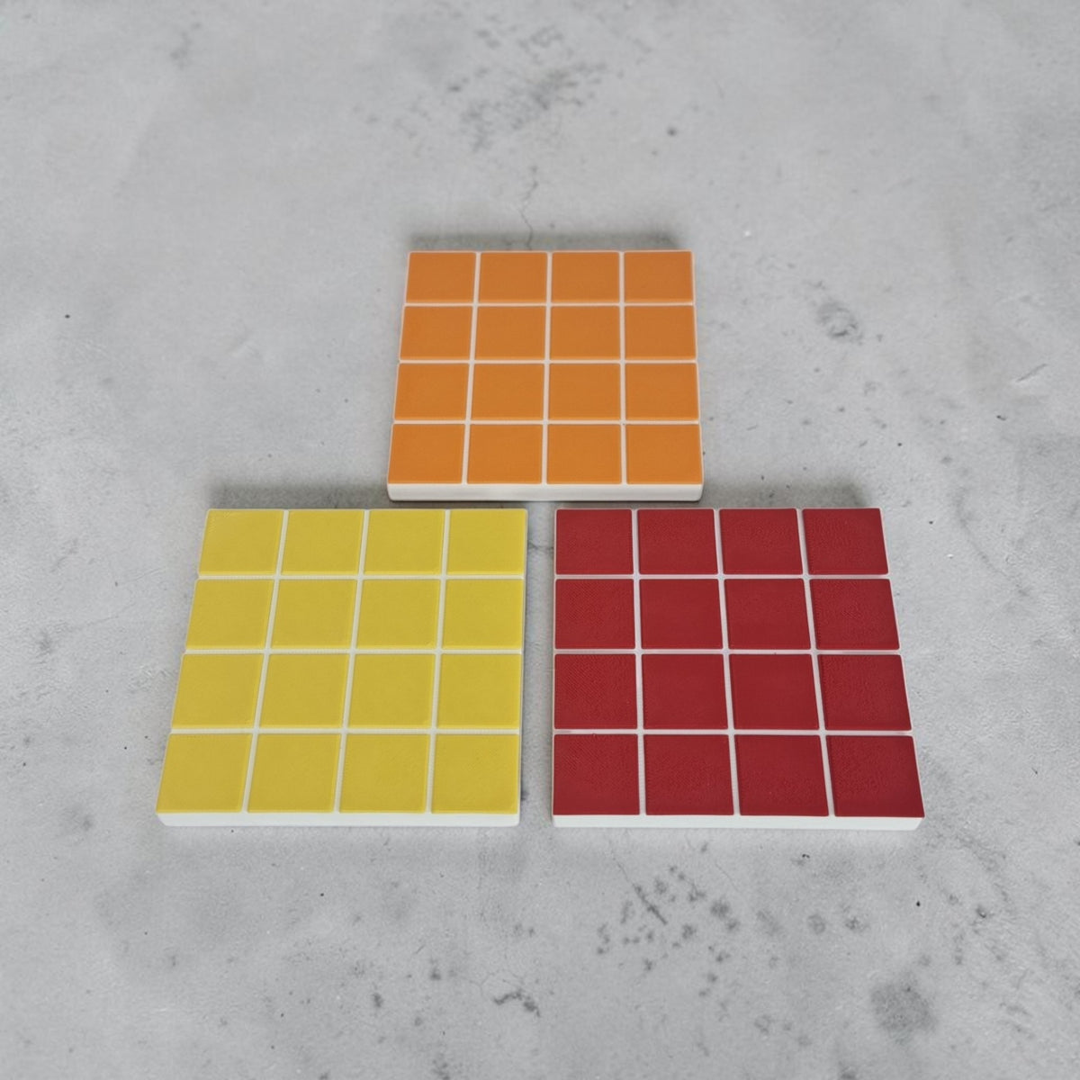 Three TILES coasters in yellow, orange and red, tile pattern with white grout lines, on a concrete surface