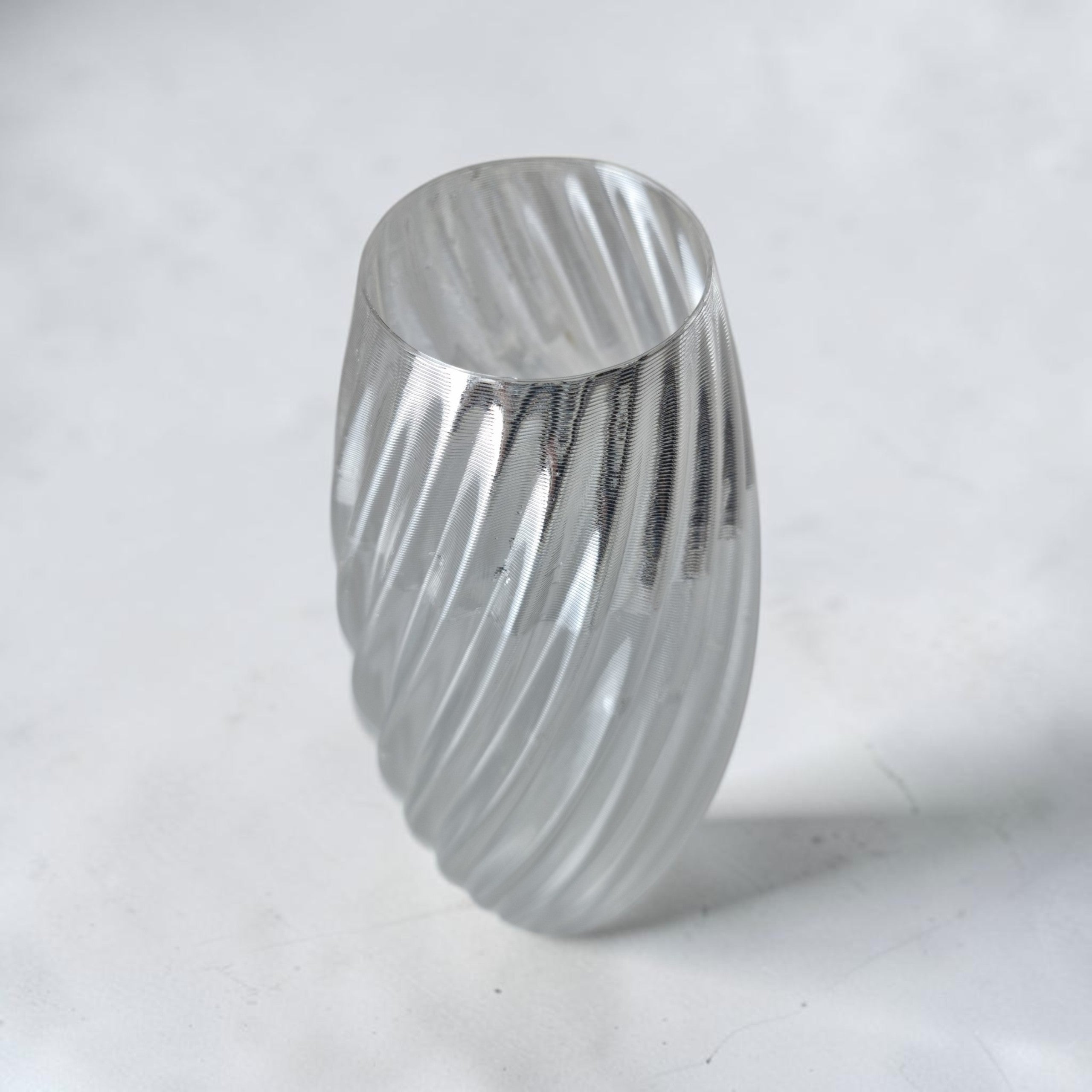 Clear spiral TEXTURIA vase in transparent bioplastic, angled view showing the twisted ribbed texture and glass-like light refraction, on a light grey studio background