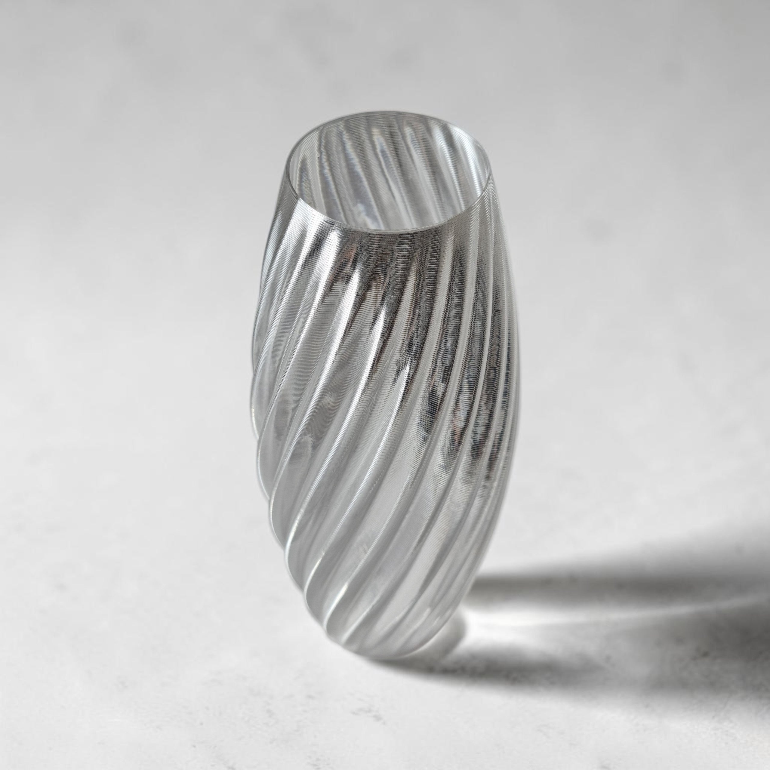 TEXTURIA Cristal vase in clear recyclable bioplastic, tilted angle with dramatic shadow, showing the full spiral silhouette and glass-effect transparency